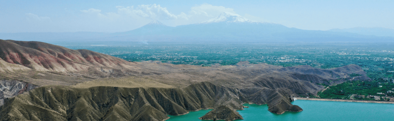 A wide, calm lake glistening in the sunlight, framed by vibrant greenery and soft hills on the horizon