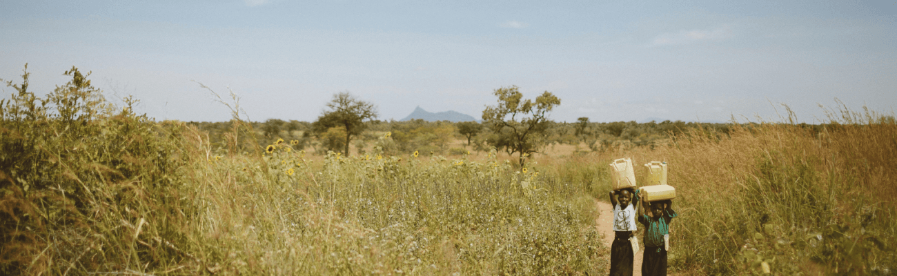 A woman carries a water container on her back while walking through a lush green field under a clear sky